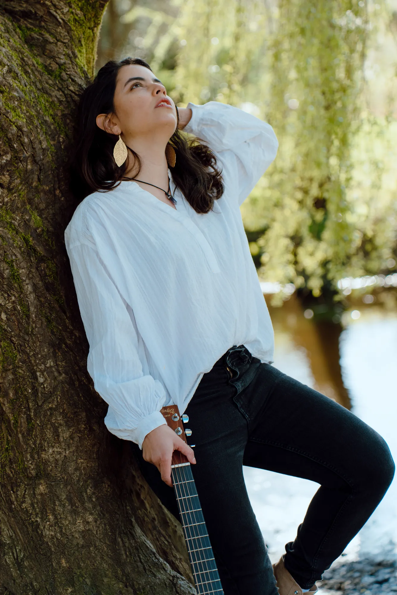 Ani leaning against the willow tree by the river, guitar in hand
