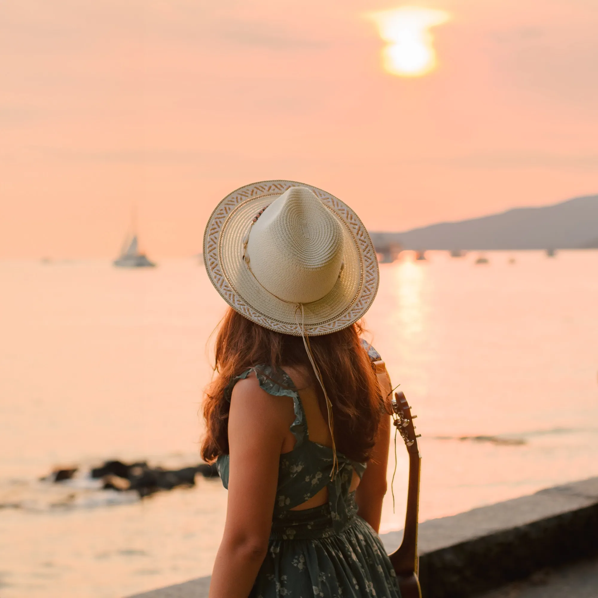 Ani in a straw hat with her ukulele, looking out at the sunset over the water