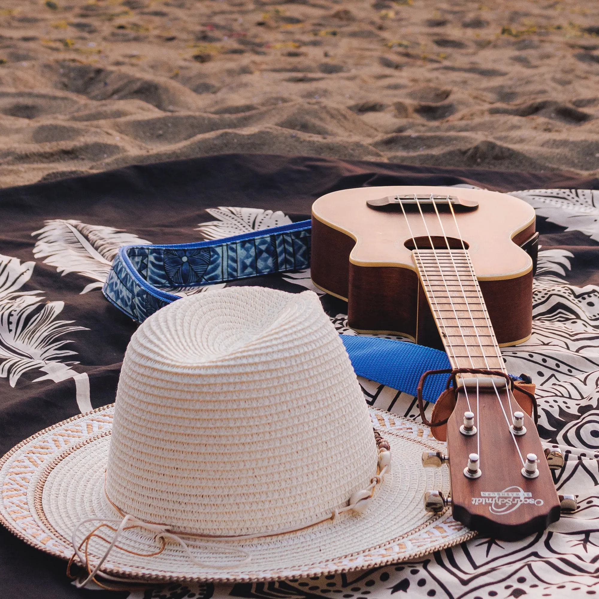 A straw hat and ukulele resting on a patterned blanket on the sand